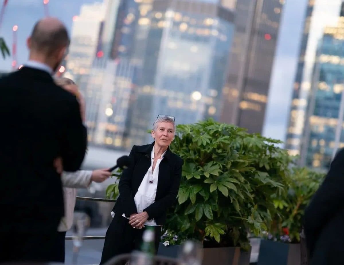 A woman in a black jacket and white shirt at an evening event, with a city skyline behind her.