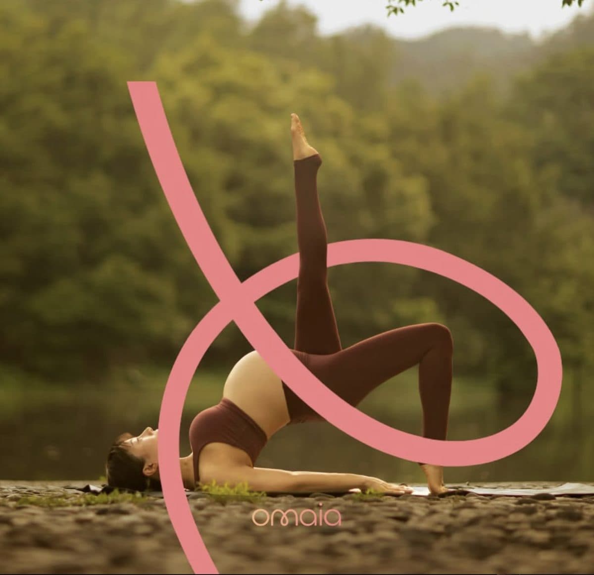 A pregnant woman in a yoga bridge pose beside a river, with the pink Omaia ribbon looping around her.